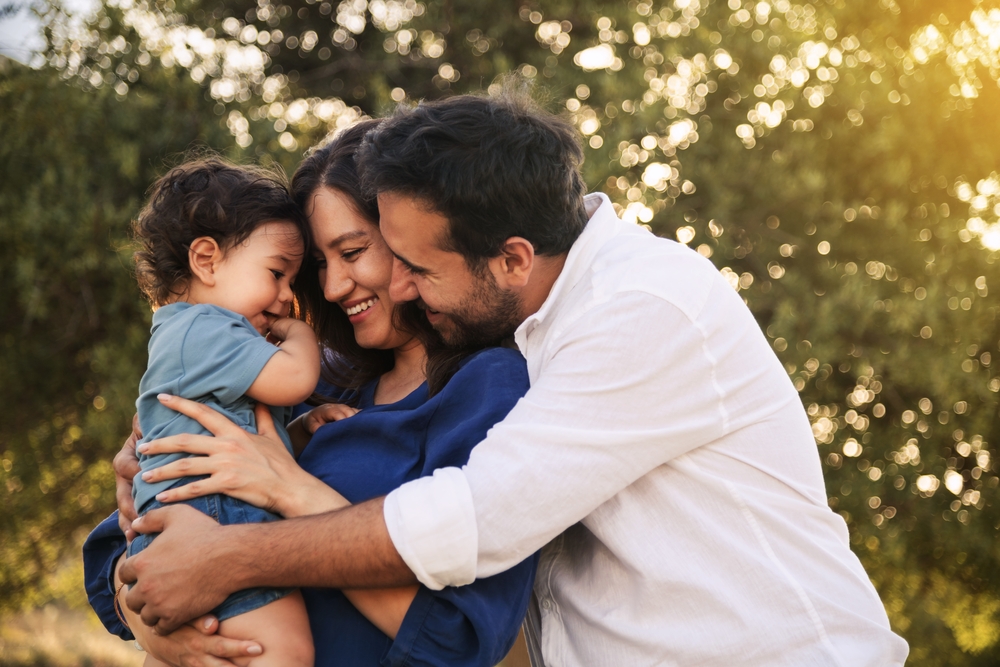 Estate Planning for Young Families with Minor Children A joyful family moment with a father, mother in a blue dress, and their one-year-old son in blue clothes, hugging and smiling in a grassy natural setting with a tree in the background.