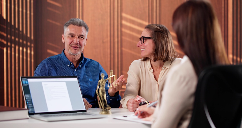 Three colleagues in a law-office meeting around a desk with a laptop and a Lady Justice statue on display.