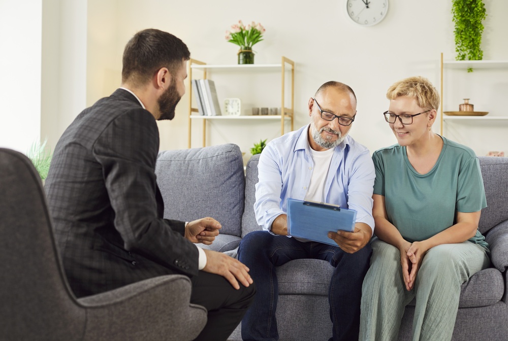 SmilingElderlyCoupleMeetsAgentAtHomeReviewingDocumentsAnd Senior couple reviewing and signing property and estate planning documents with a financial advisor during a home consultation.
